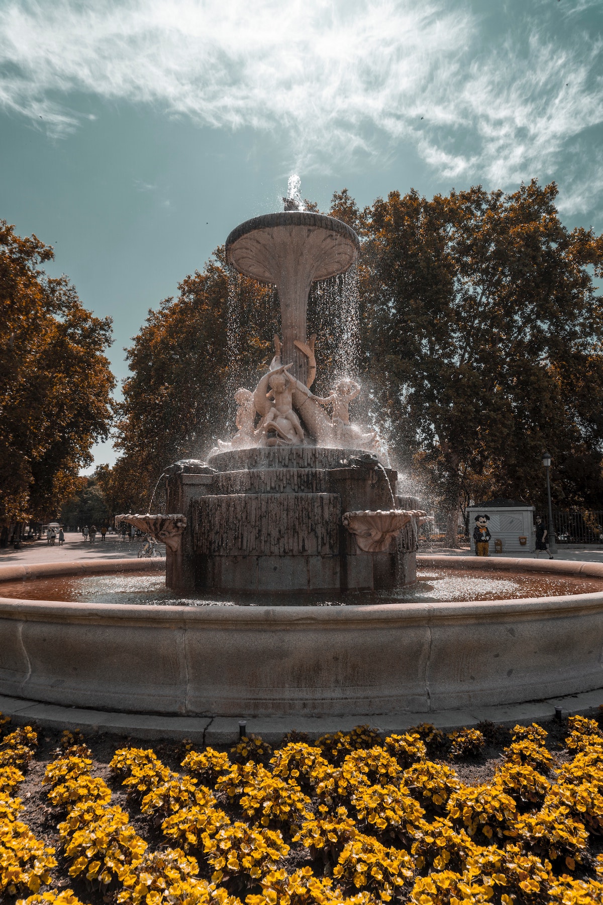 Large stone fountain in a park surrounded by yellow flowers.