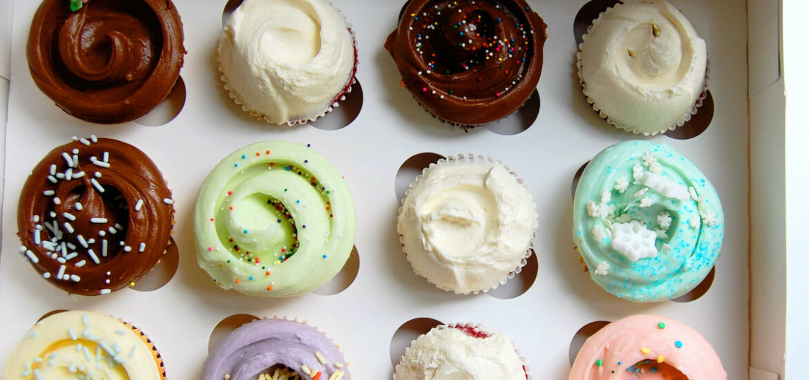Overhead shot of a dozen differently colored cupcakes in a white bakery box