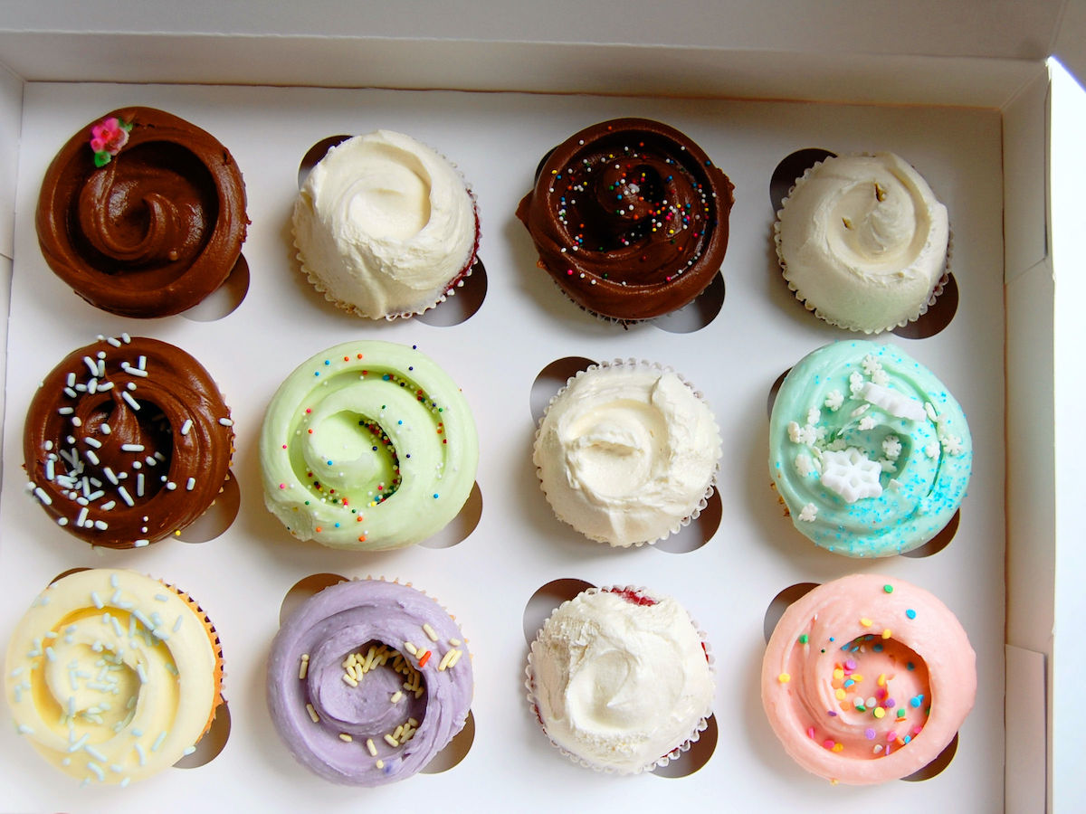 Overhead shot of a dozen differently colored cupcakes in a white bakery box