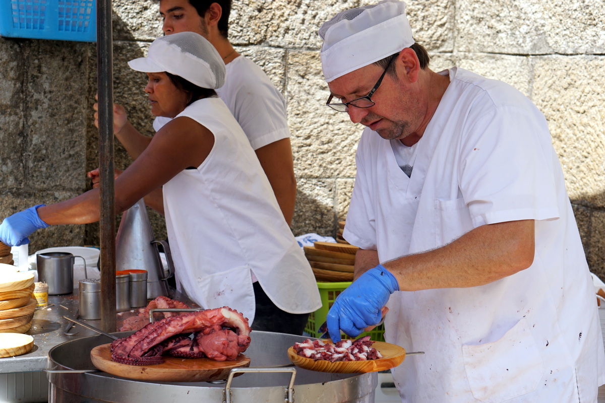 A man in a white cook's outfit cuts pieces of boiled Galician octopus onto a wooden plate.