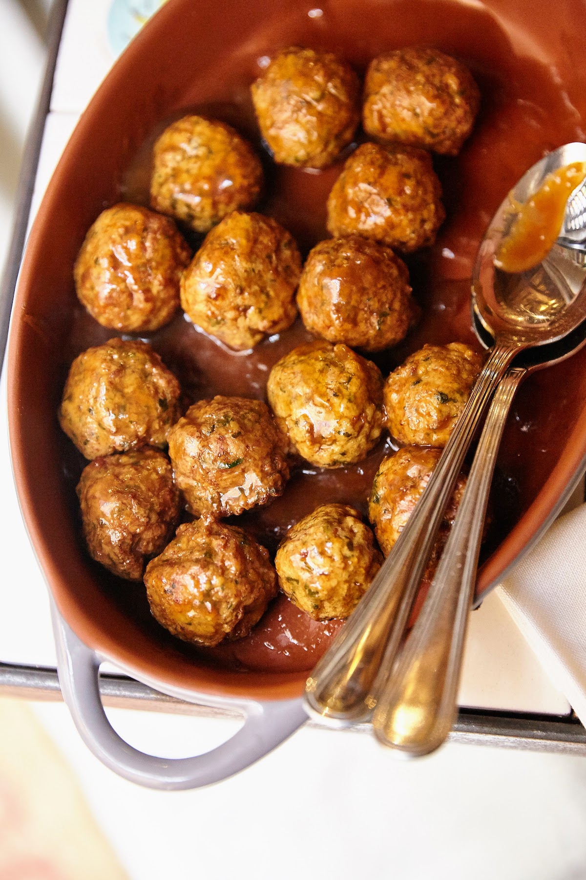 Overhead shot of meatballs and two metal spoons in a reddish-brown oval dish.