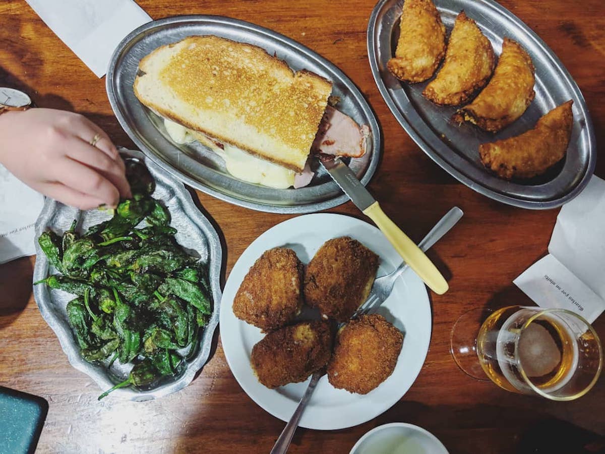 Overhead shot of a plate of fried green peppers, a large sandwich, fried croquettes, a small tray of mini empanadas, and a glass of beer.