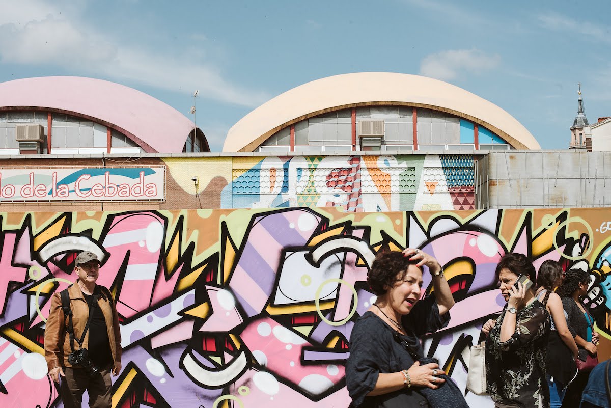 People walking in front of a colorful street art mural on the exterior walls of a large market building.