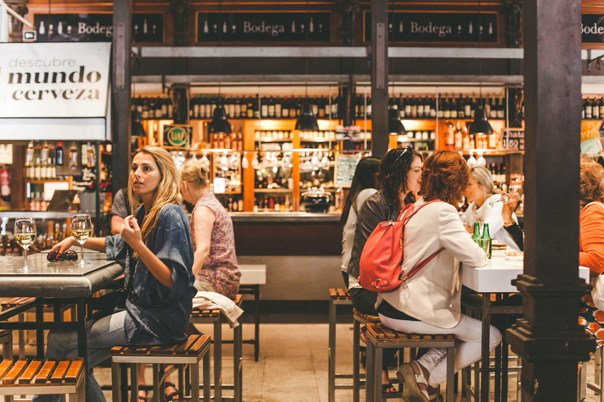 People seated at high tables inside a food hall eating tapas and drinking wine