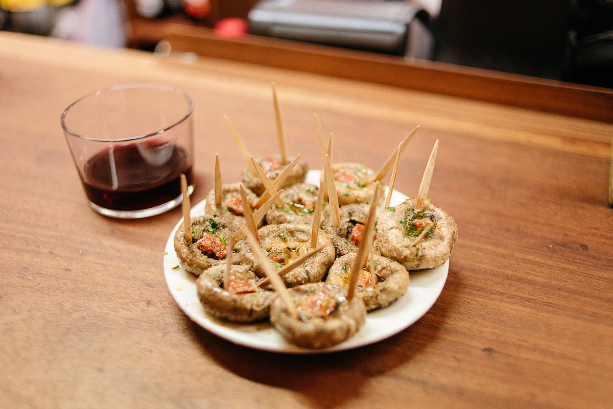 Plate of grilled mushrooms with chorizo next to a short glass of red wine.