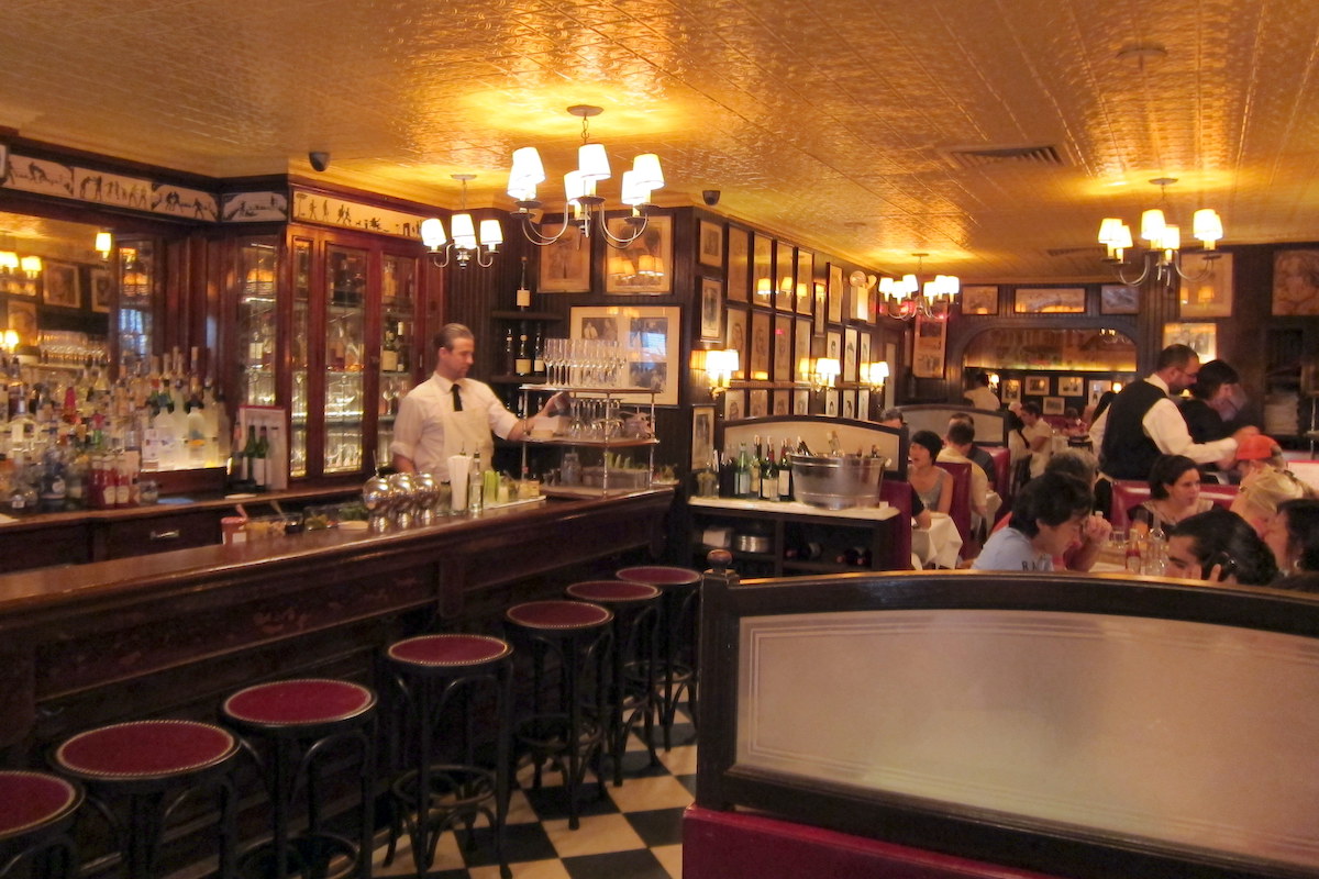 Old fashioned bar with checkered floor tiles, dark wood paneling, and rows of framed pictures on the wall