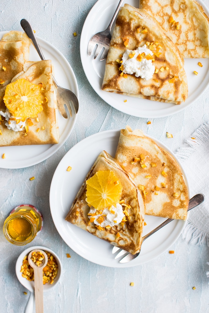 Three plates each with two crepes, topped with orange slices, honey, whipped cream and nuts
