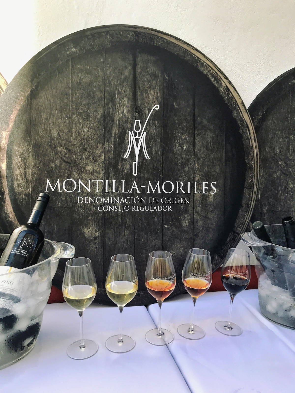 Five glasses of wine, ranging in color from pale yellow to dark brown, on a white table top in front of a black barrel reading "Montilla-Moriles."