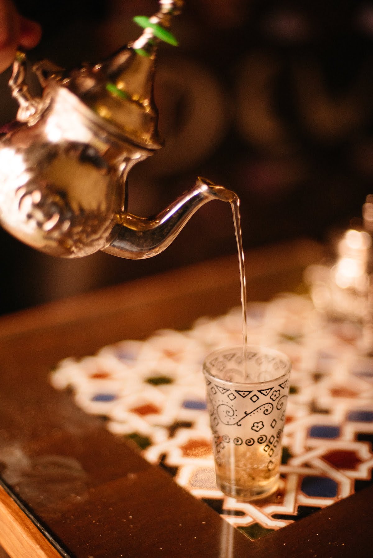 Moroccan tea being poured from a metal teapot into a handleless glass cup on a tiled table.