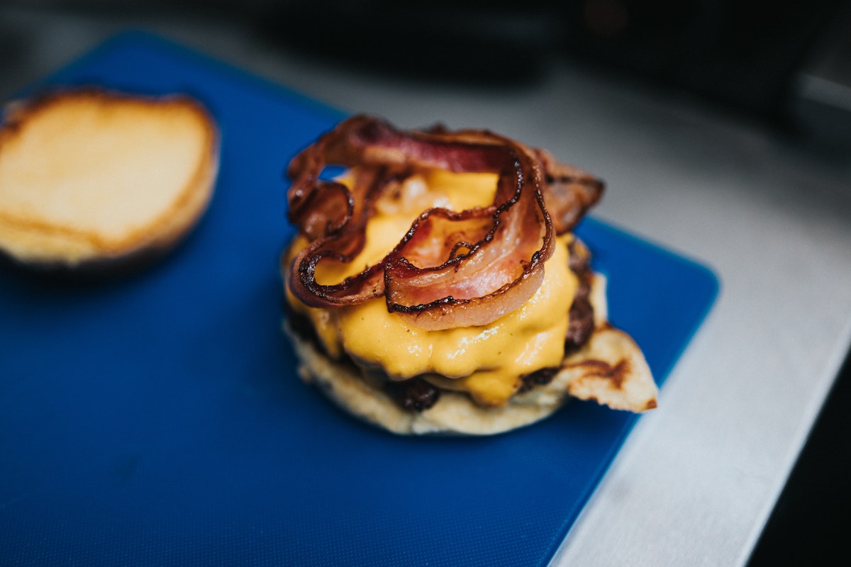 A bacon cheese burger sits on a blue cutting board with the top bun on the side so we can see the crispy bacon and yellow melty cheese 