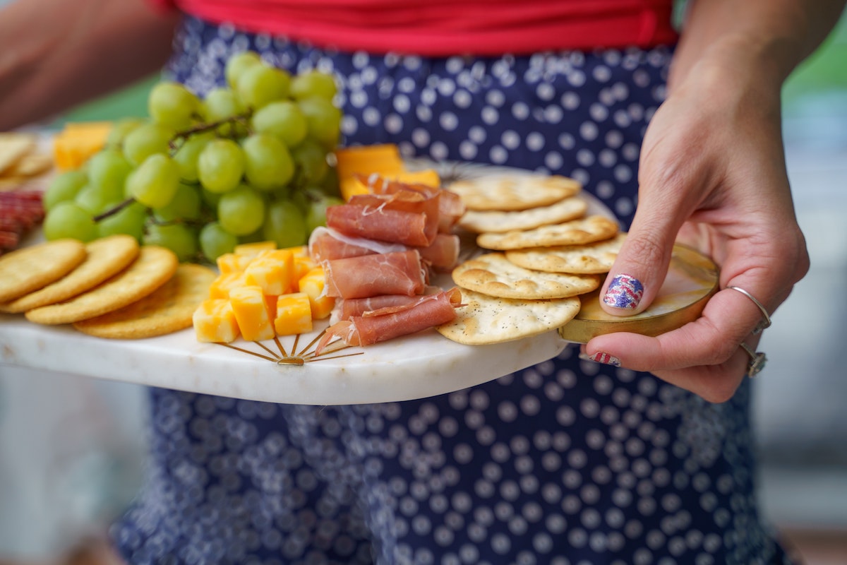 person holding a white platter covered with cheese, cold cuts, crackers, and fruit