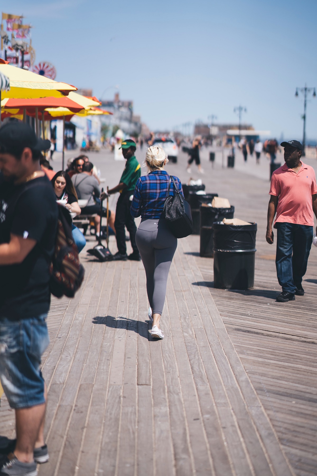 Young woman with large bag walks down a boardwalk with many people in the hot sun