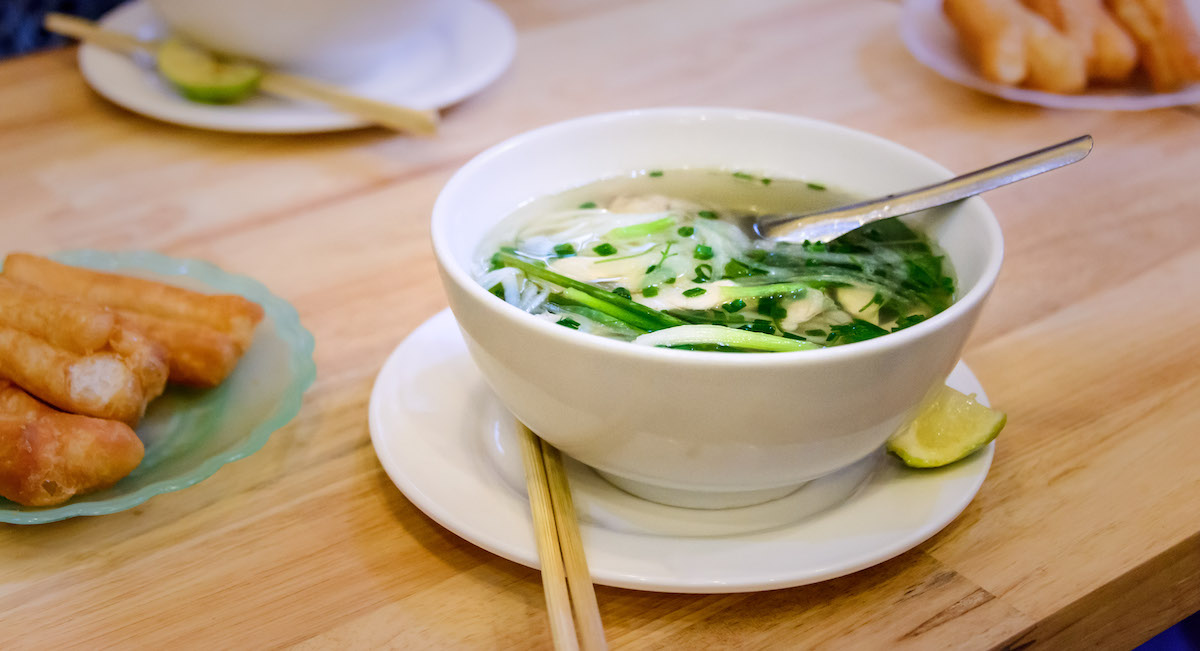 Bowl of noodle soup with chives and green vegetables on a white plate beside a citrus wedge and a pair of chopsticks