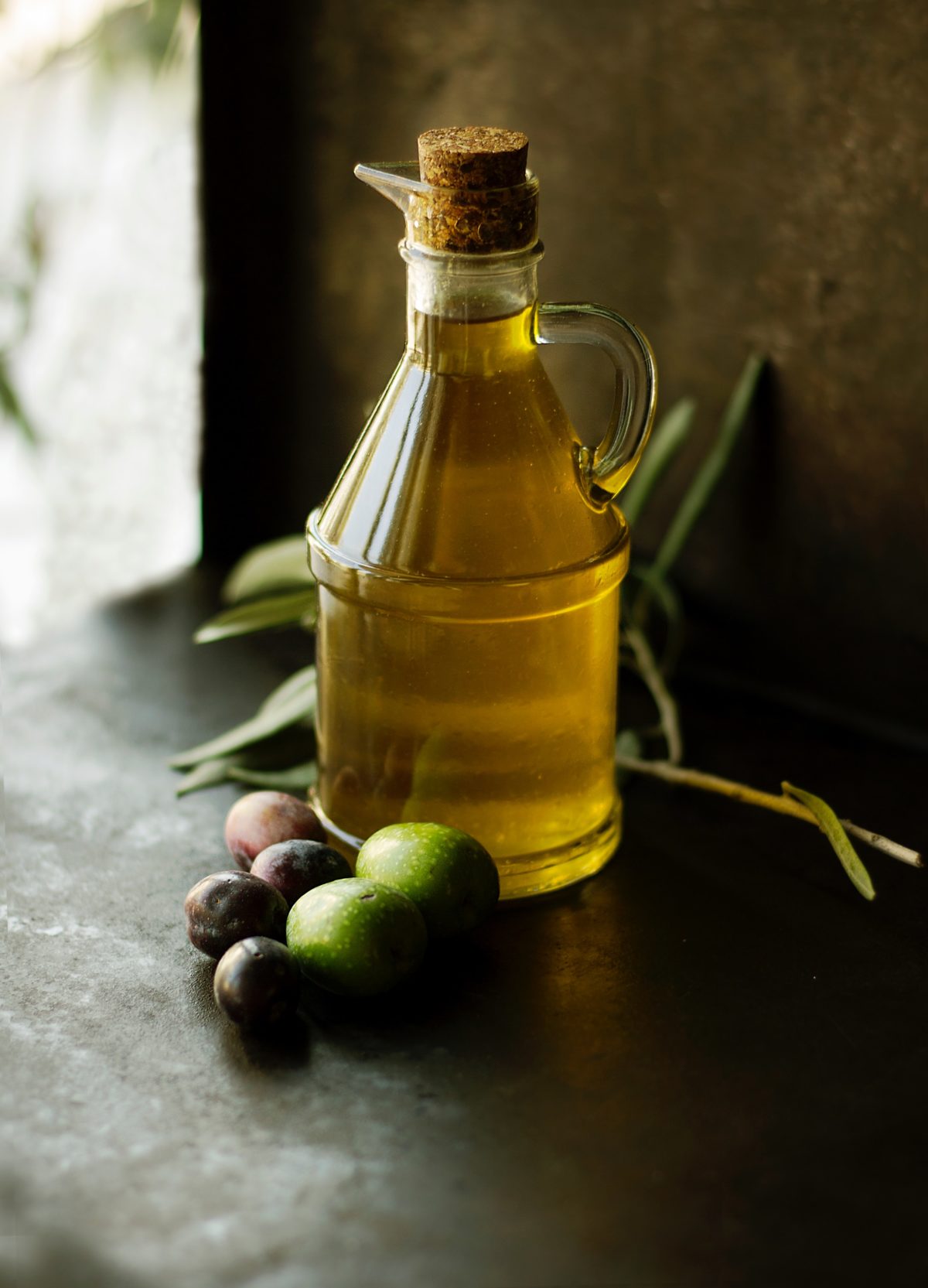 A clear glass bottle of olive oil on a dark tabletop next to a handful of olives.