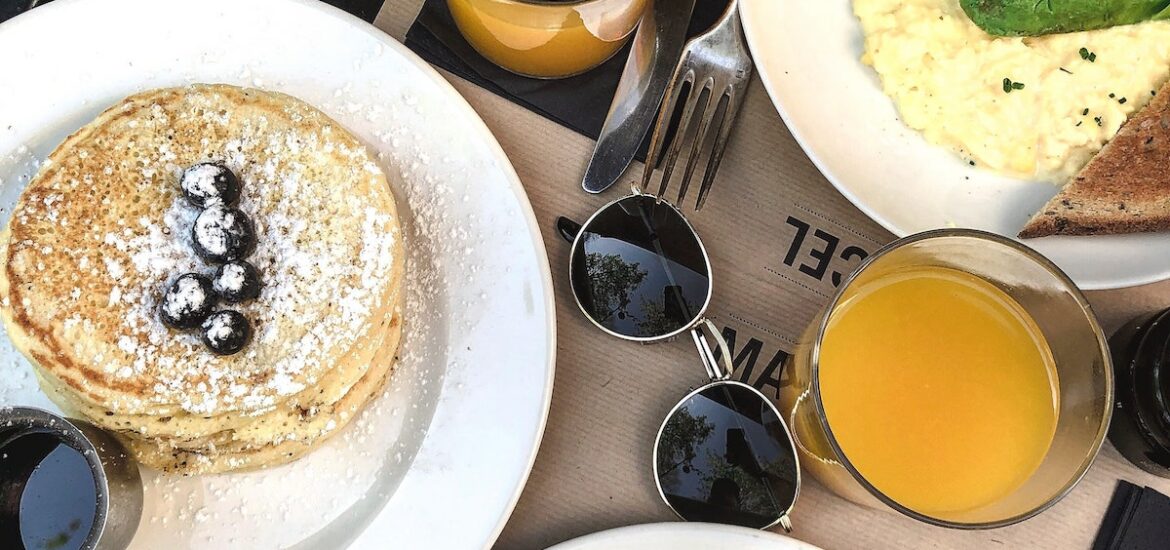 Overhead shot of breakfast items on a table including pancakes, eggs Benedict, toast, and two glasses of orange juice, with a pair of sunglasses in the center