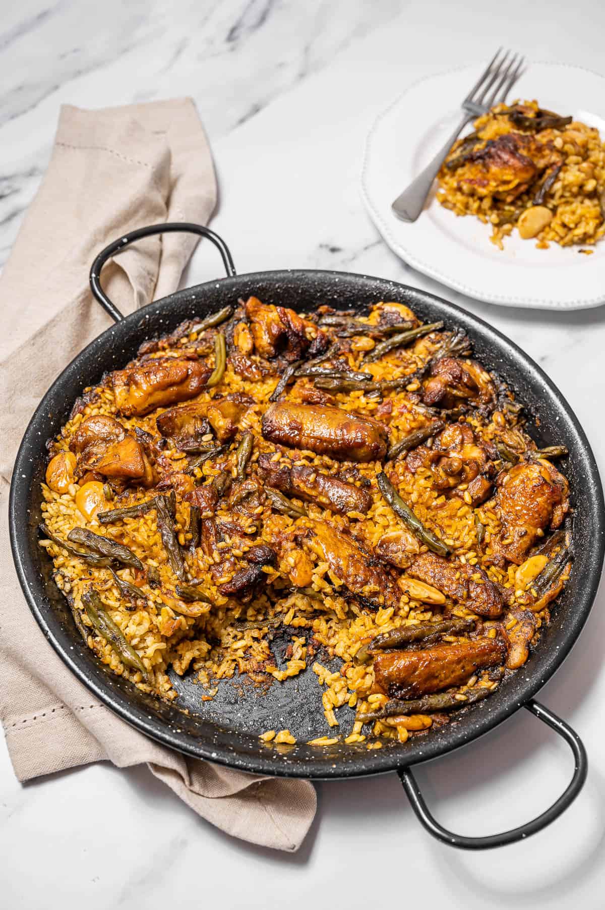 A large pan of Paella Valenciana with one portion plated behind it.