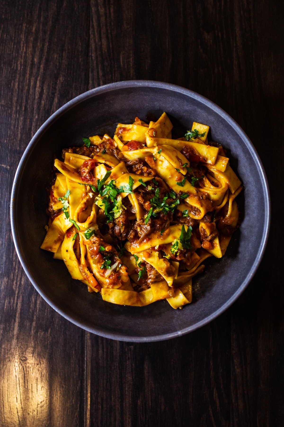 Overhead shot of long, wide pasta with meat sauce and herbs in a bowl against a dark background