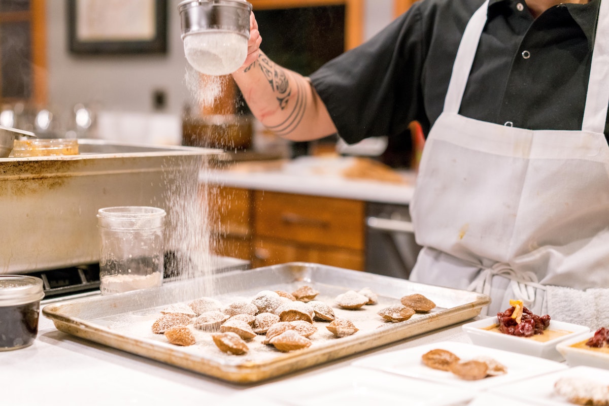 A pastry chef wearing a black shirt and white apron sprinkling powdered sugar over small food items on a baking tray