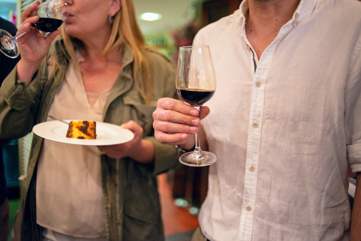 Person holding a glass of dark sherry wine next to another person holding a plate of cheesecake and drinking wine.