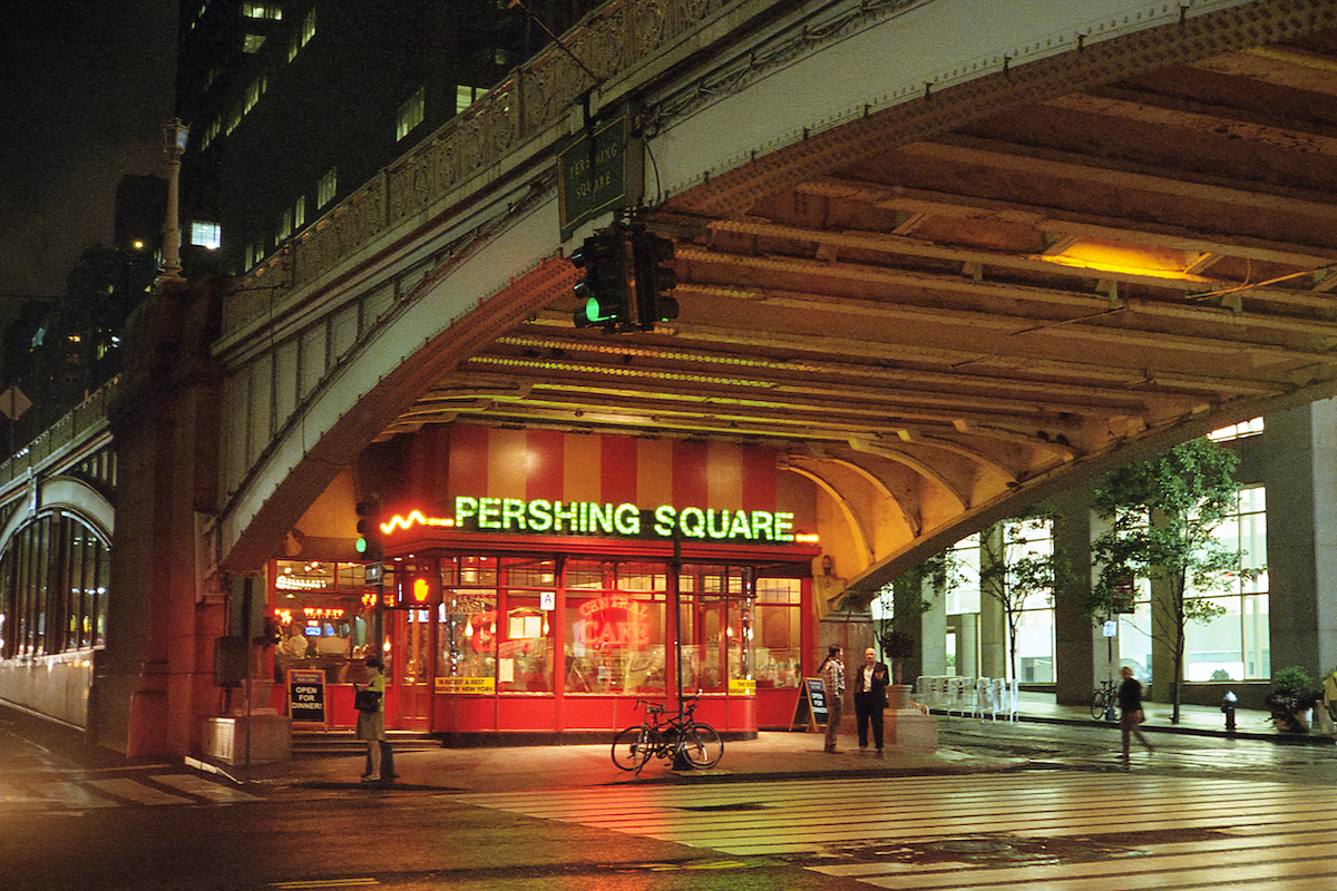 Brightly lit diner with green fluorescent letters reading Pershing Square under an overpass in an urban area