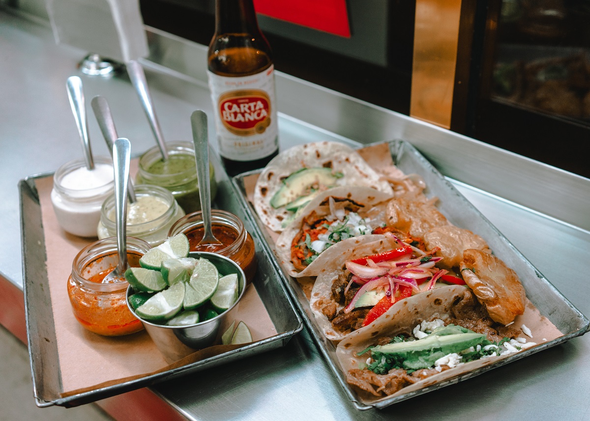 A tray of street tacos with limes, sauces, and beer