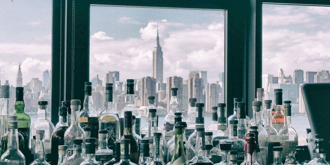 A full counter of bottles of spirits in front of a window looking out at the Manhattan skyline