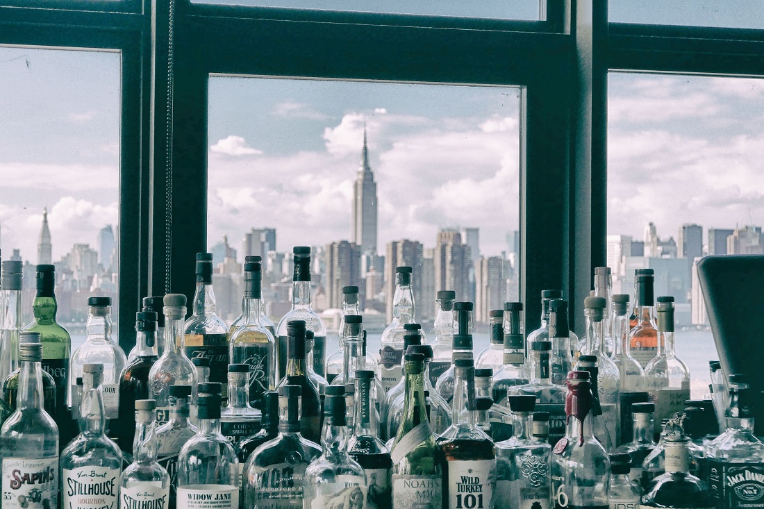 A full counter of bottles of spirits in front of a window looking out at the Manhattan skyline