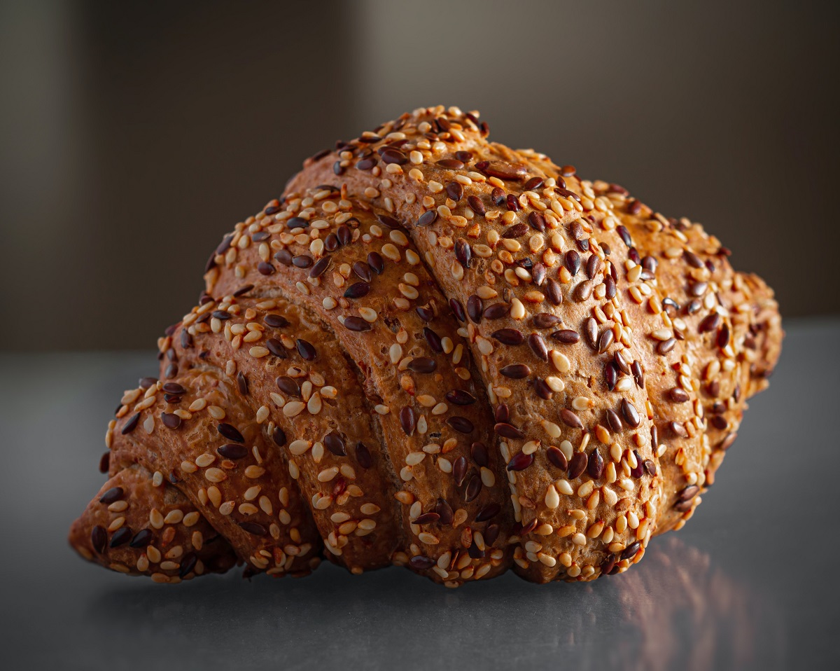 Plump croissant covered in different colored seeds on top of a reflective surface