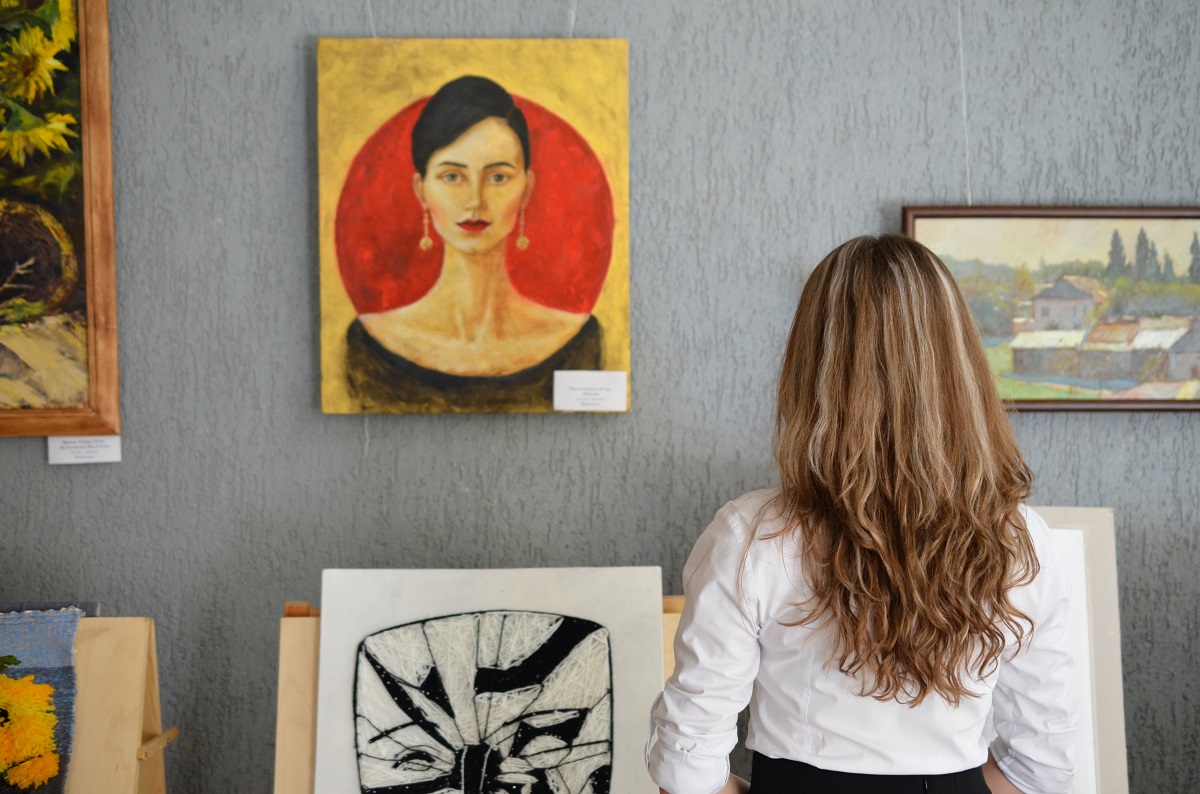 A woman in a white shirt looks at a portrait painting of a serious woman