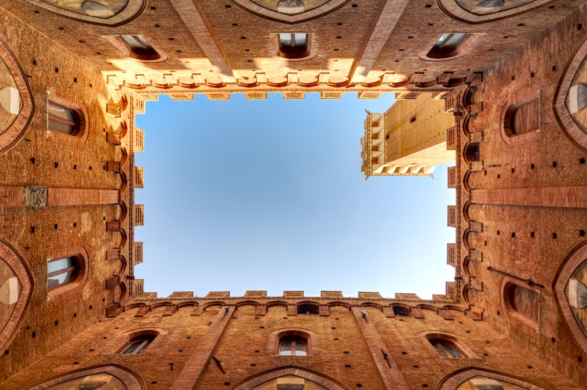 View of the sky taken from within a rectangular courtyard with brown medieval buildings and a tower visible around the edges.