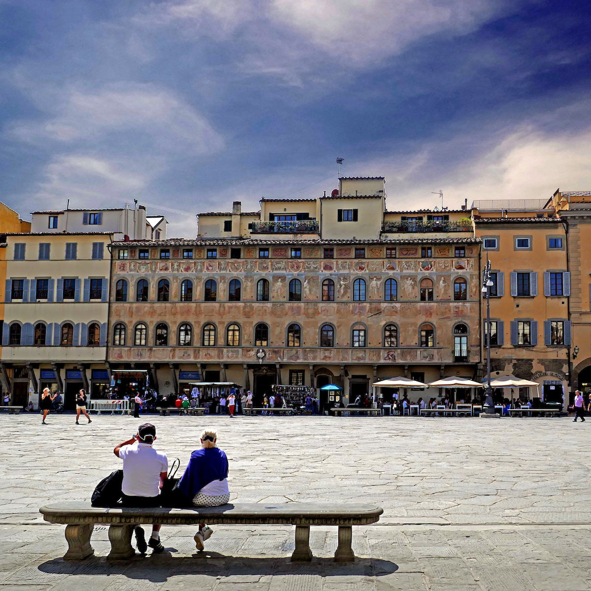 Large open square in Florence, Italy with two people sitting on a stone bench in the foreground.