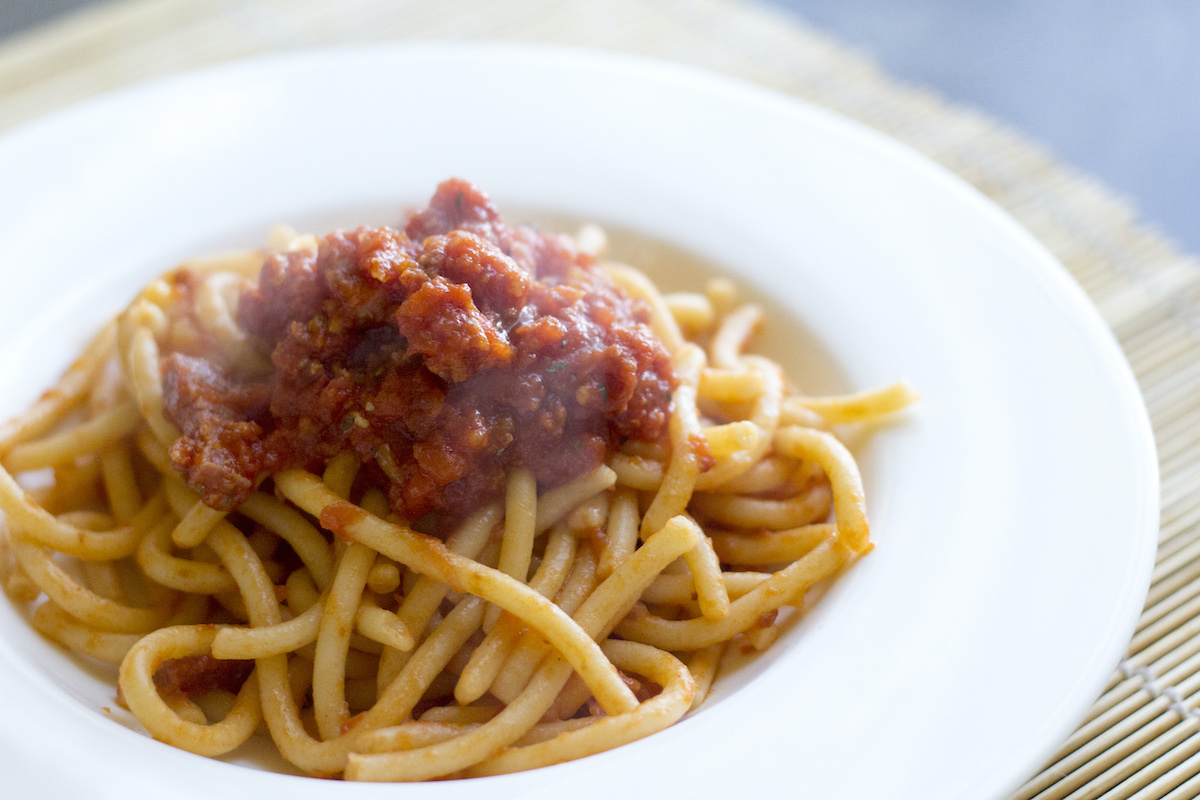 Thick spaghetti-like noodles covered in a sausage-based sauce, served on a white plate