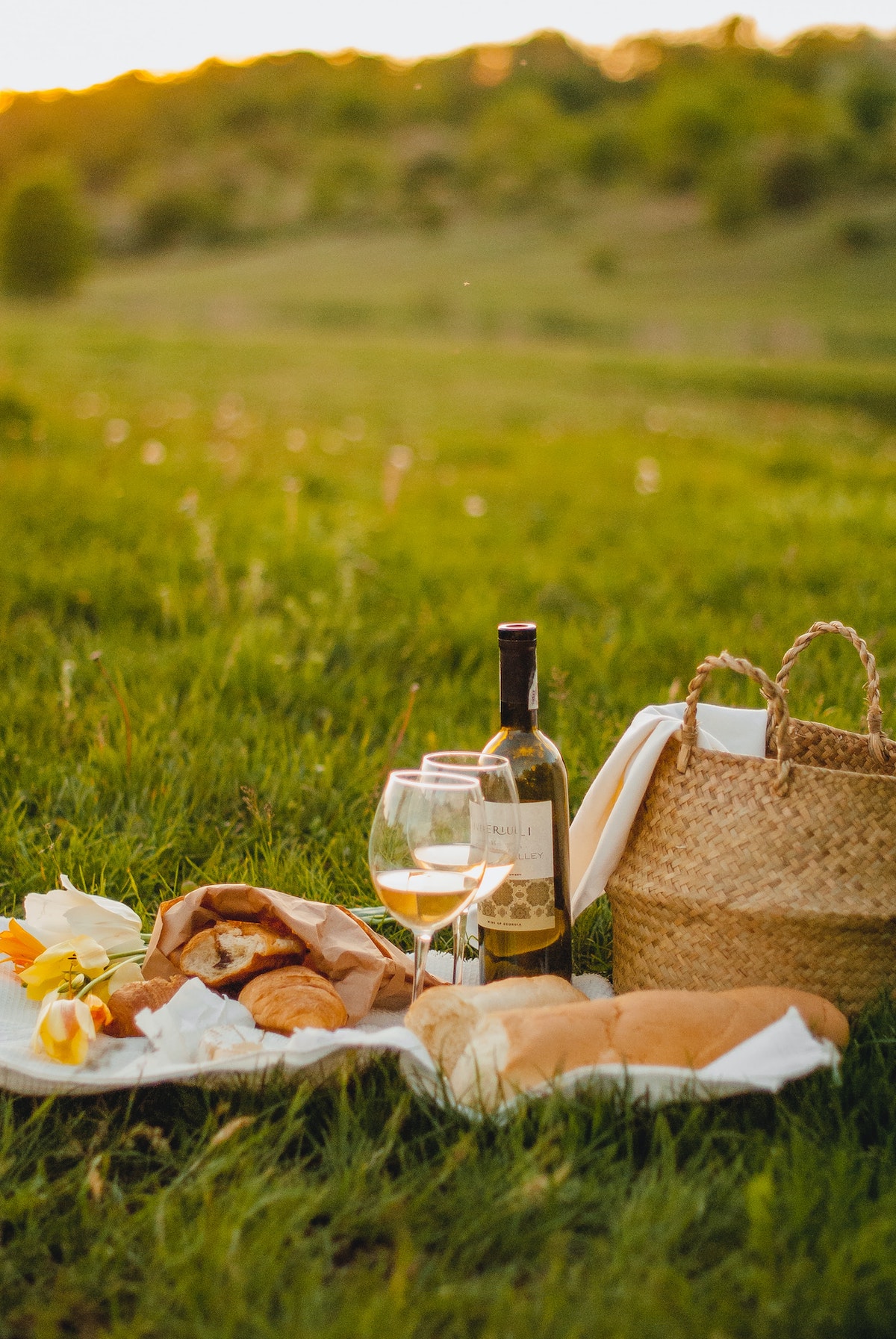 Picnic spread of bread, pastries, and wine next to a brown wicker tote in a grassy field.