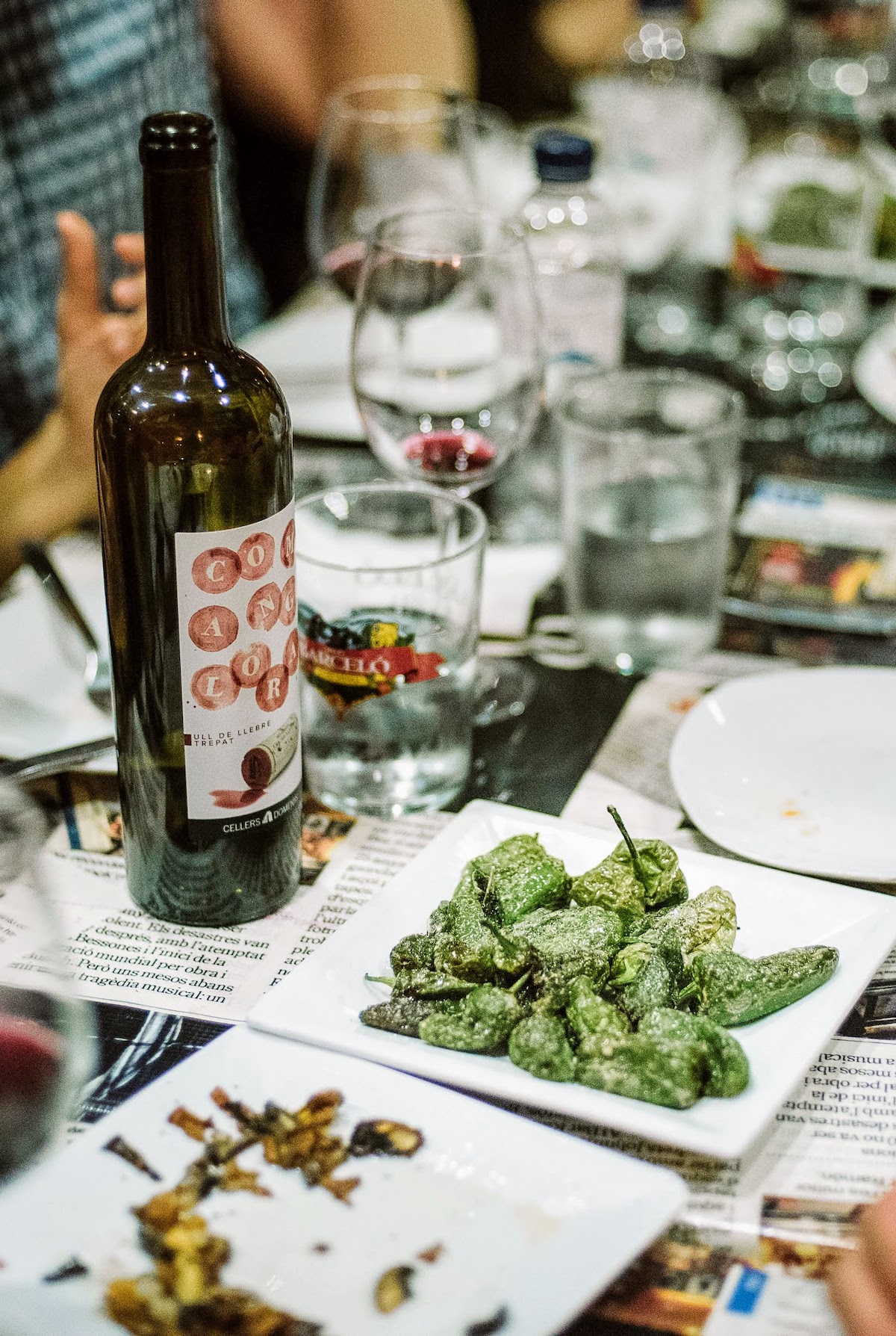 Small green peppers on a white plate beside a bottle of wine on a crowded tabletop