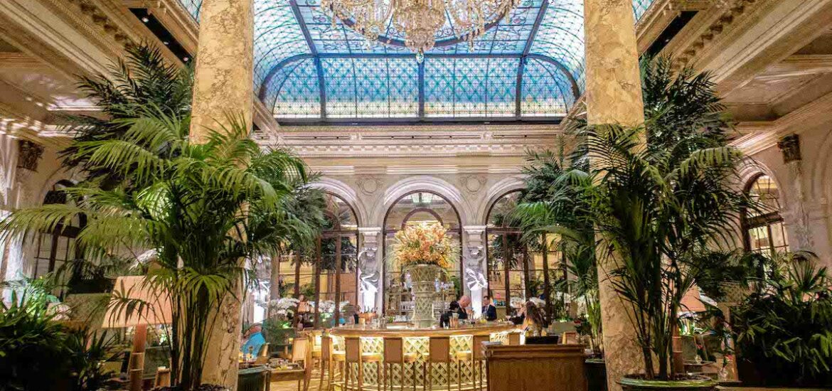 Ornate luxury hotel bar with large chandelier overhead and entrance flanked by potted plants and Greek style columns