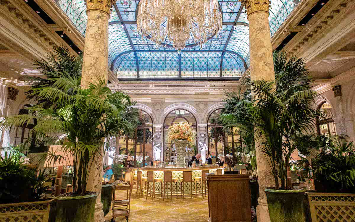 Ornate luxury hotel bar with large chandelier overhead and entrance flanked by potted plants and Greek style columns