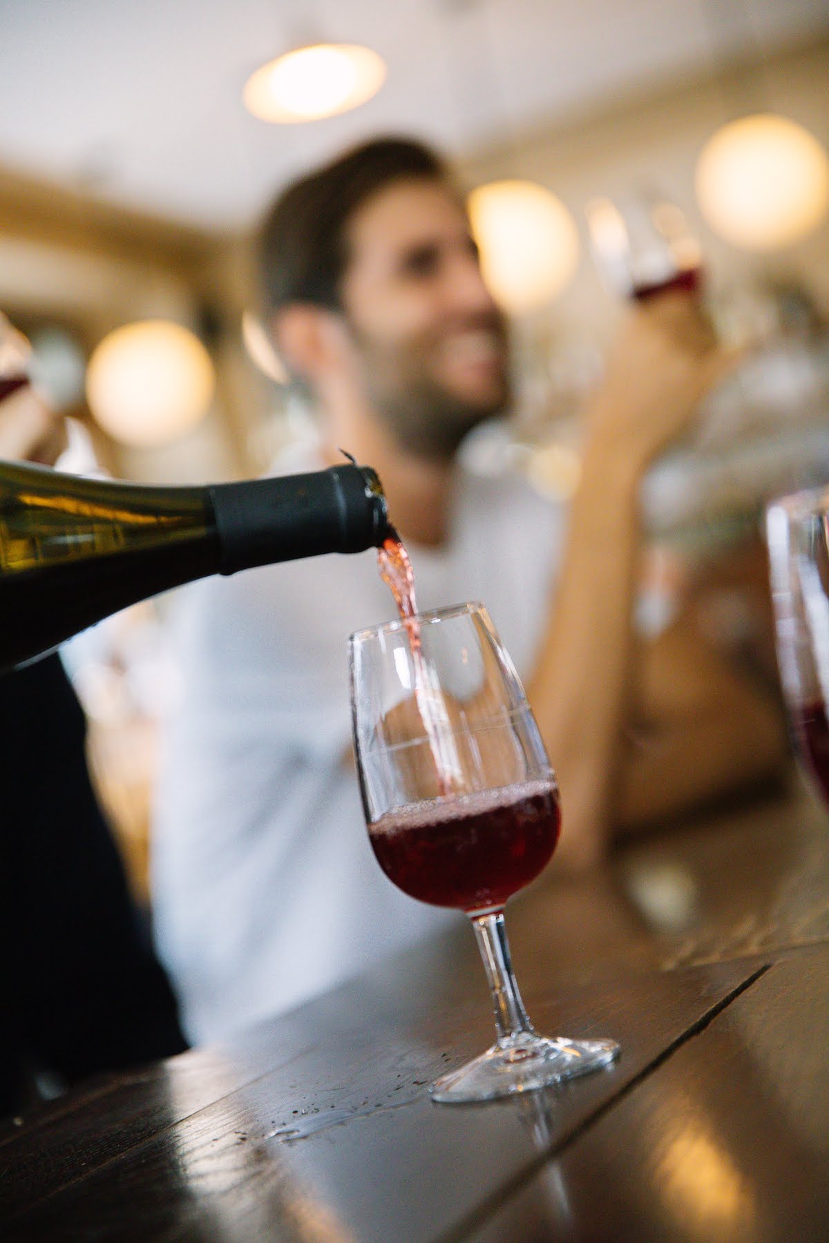 Close up of a small glass of red wine being poured with a smiling man holding another glass of wine in the background