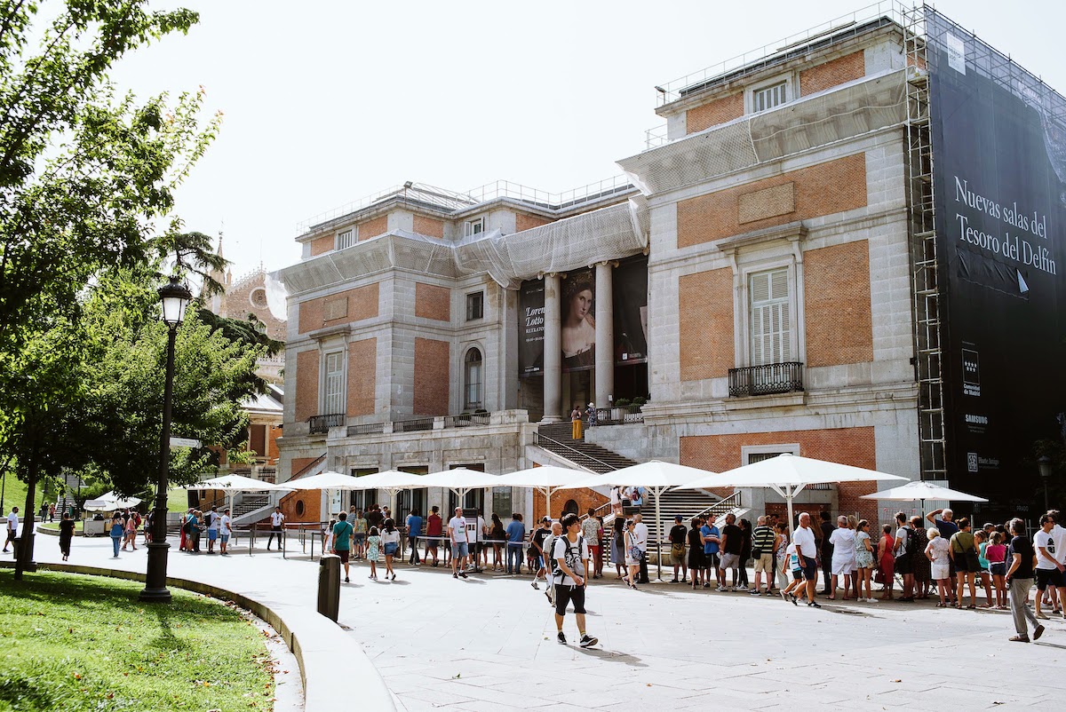 Exterior of a large brick and stone building with people standing in line under shady umbrellas outside.