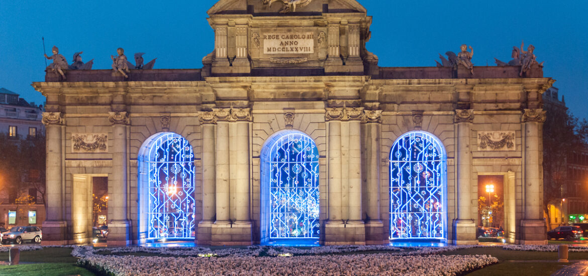Large stone gateway decorated with blue holiday lights with pink and white flowers in front.