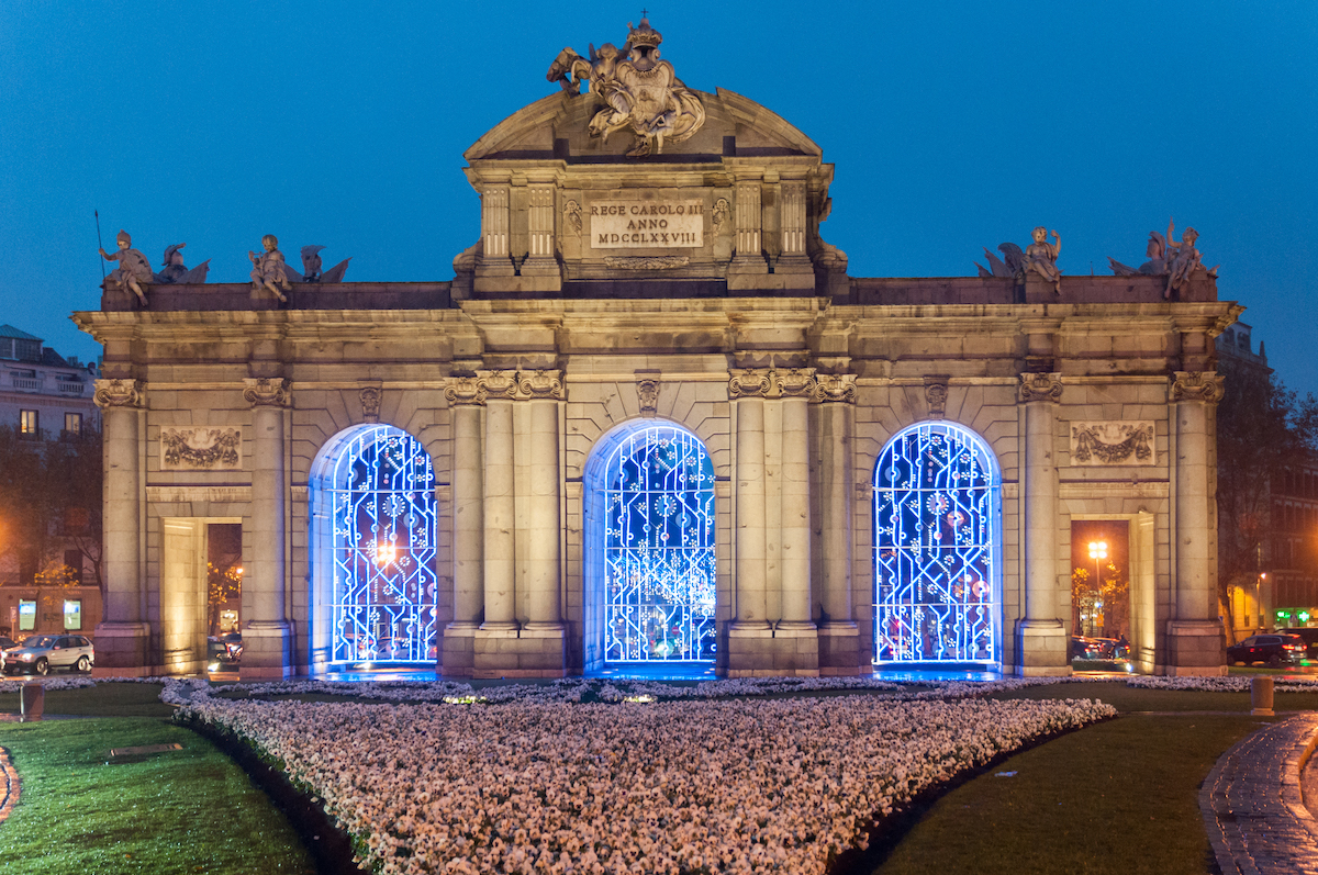 Large stone gateway decorated with blue holiday lights with pink and white flowers in front.