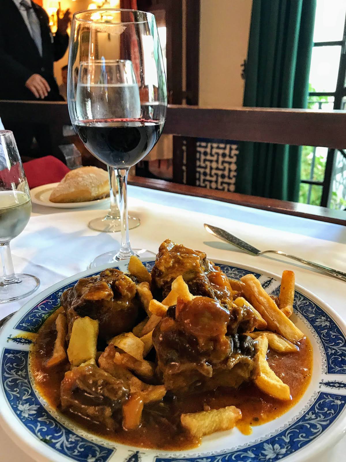 Plate of bull tail stew with french fries next to a glass of red wine.