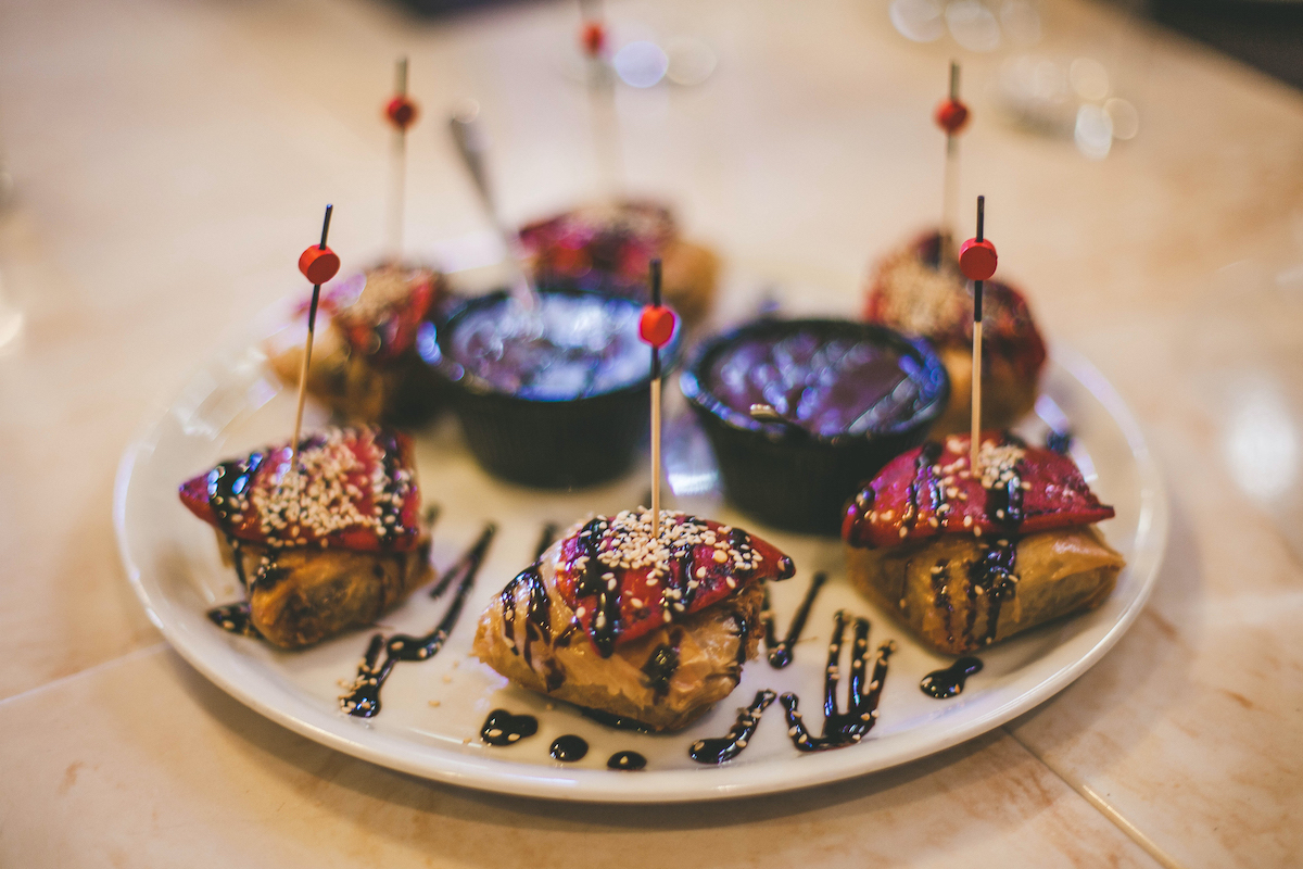 Savory pastries with roasted red peppers skewered on top arranged on a white plate with two dishes of brown sauce in the middle