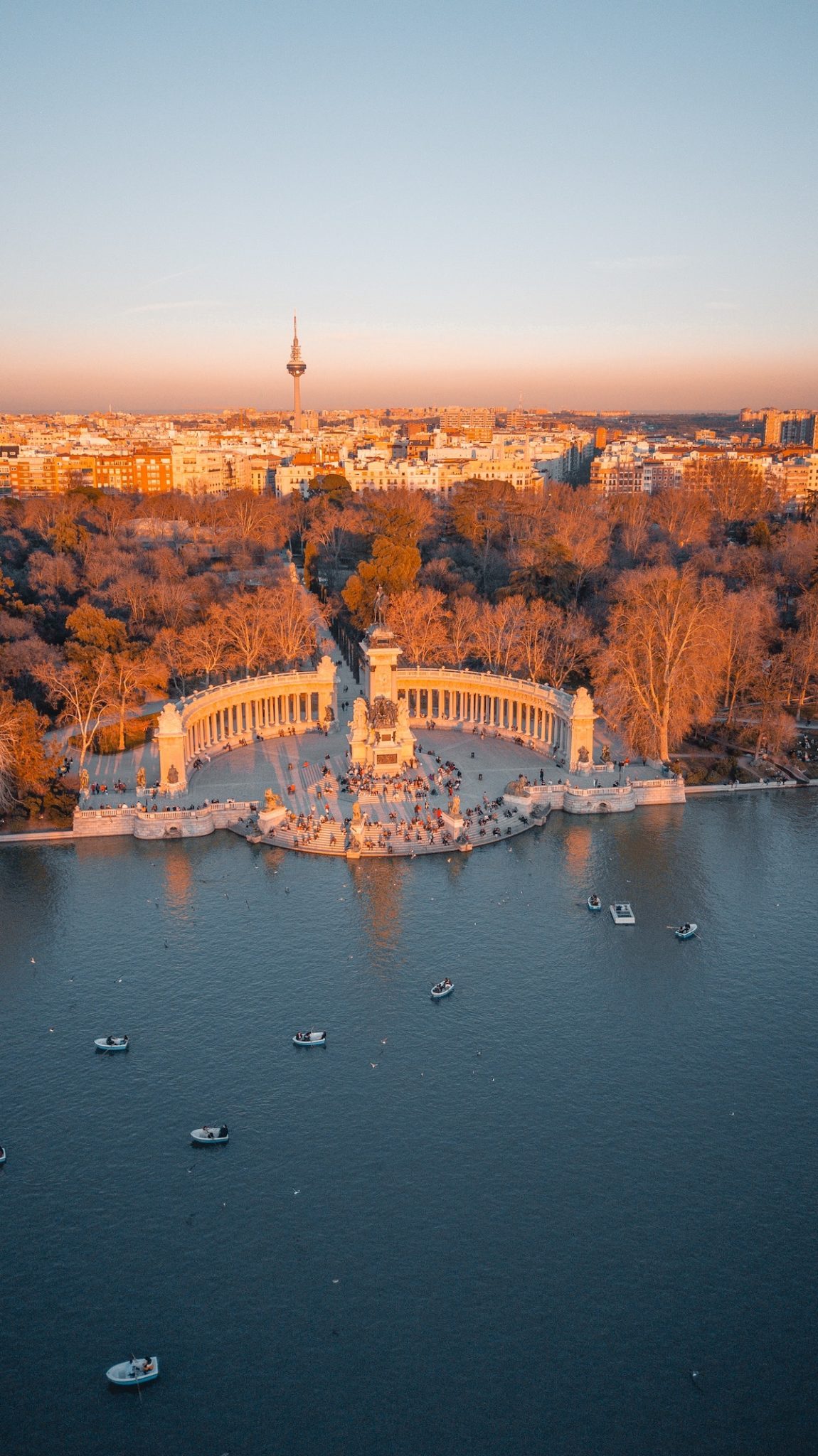 Bird's eye view of the lake in Madrid's Retiro Park, surrounded by a large stone monument and many trees with the city skyline in the background.