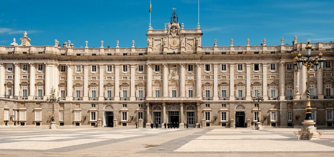 Exterior of a large, grandiose royal palace as seen from across an expansive plaza on a clear day.