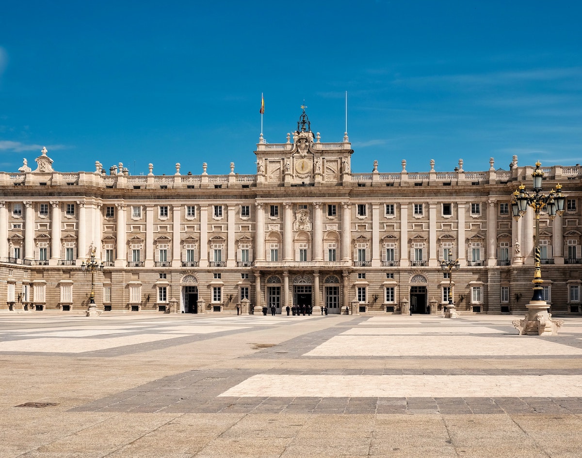 Exterior of a large, grandiose royal palace in Madrid as seen from across an expansive plaza on a clear day.
