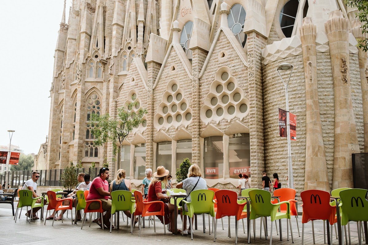 People eating outside on the terrace of a fast food restaurant in front of a large stone church.