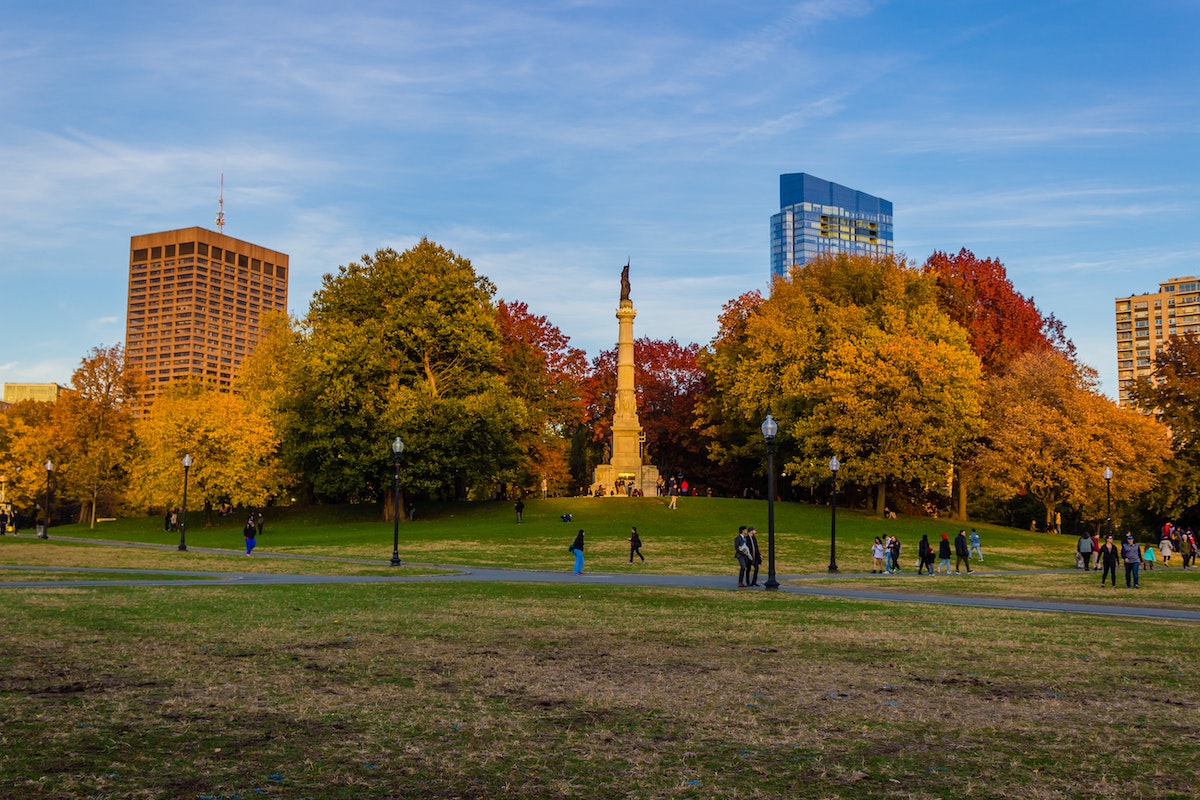 The fall landscape of Boston Public Garden. People walk along paved paths, with vibrantly colored trees all around. Skyscrapers peek out in the background.