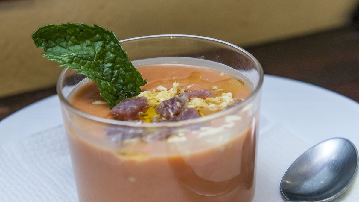 Close up of a glass of chilled tomato soup garnished with cured ham, olive oil, hard boiled egg, and a fresh parsley leaf.