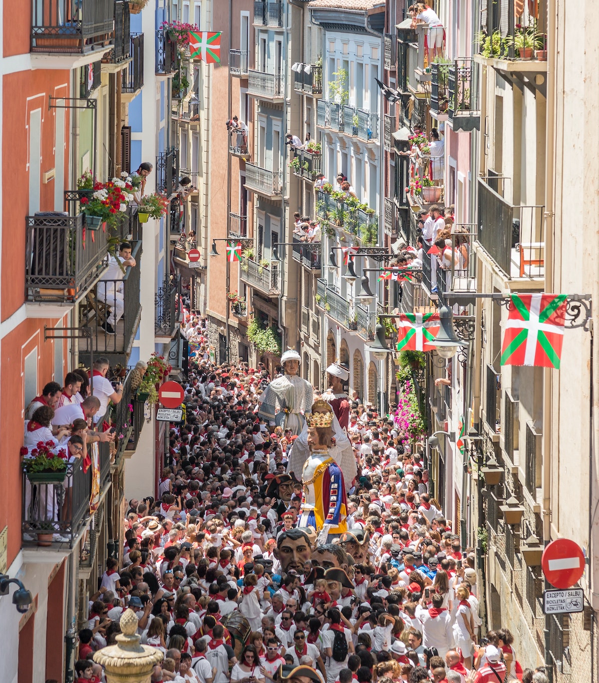 Large figurines being paraded through a crowded city street in Spain while people watch from their balconies.