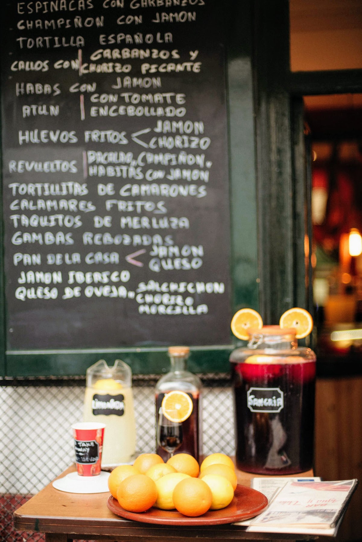 Large jugs of sangria and lemonade behind a tray of oranges, with a chalkboard menu in the background.